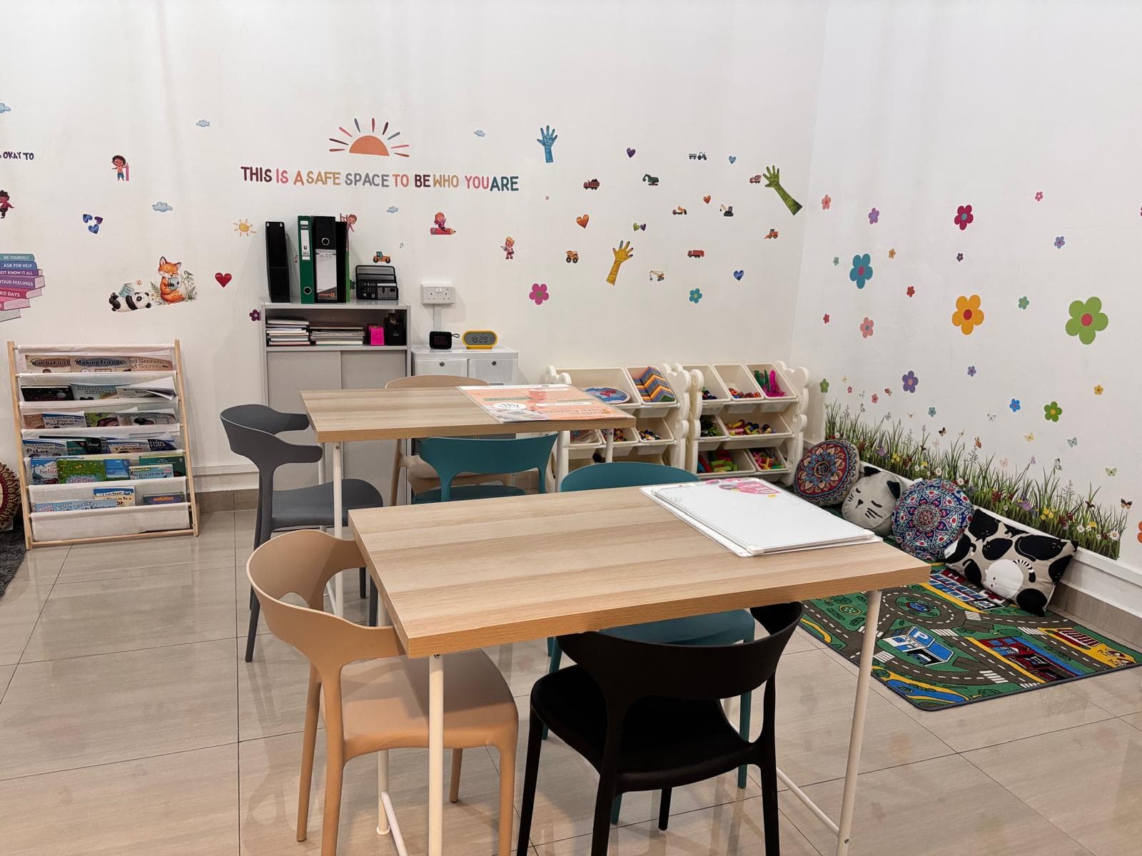 The My Skool Home room — study tables under a wall that reads “This is a safe space to be who you are”, with a bookshelf, toy shelves and a play mat